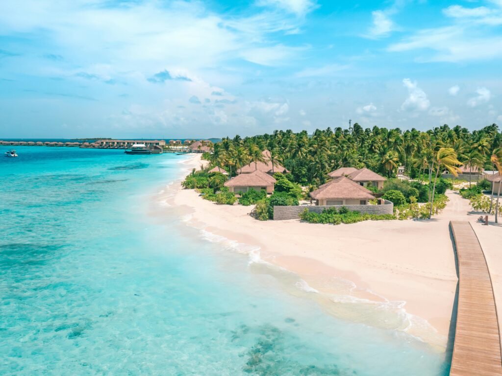 From above of pure ocean with sandy shore near wooden boardwalk and trees with small houses under blue cloudy sky